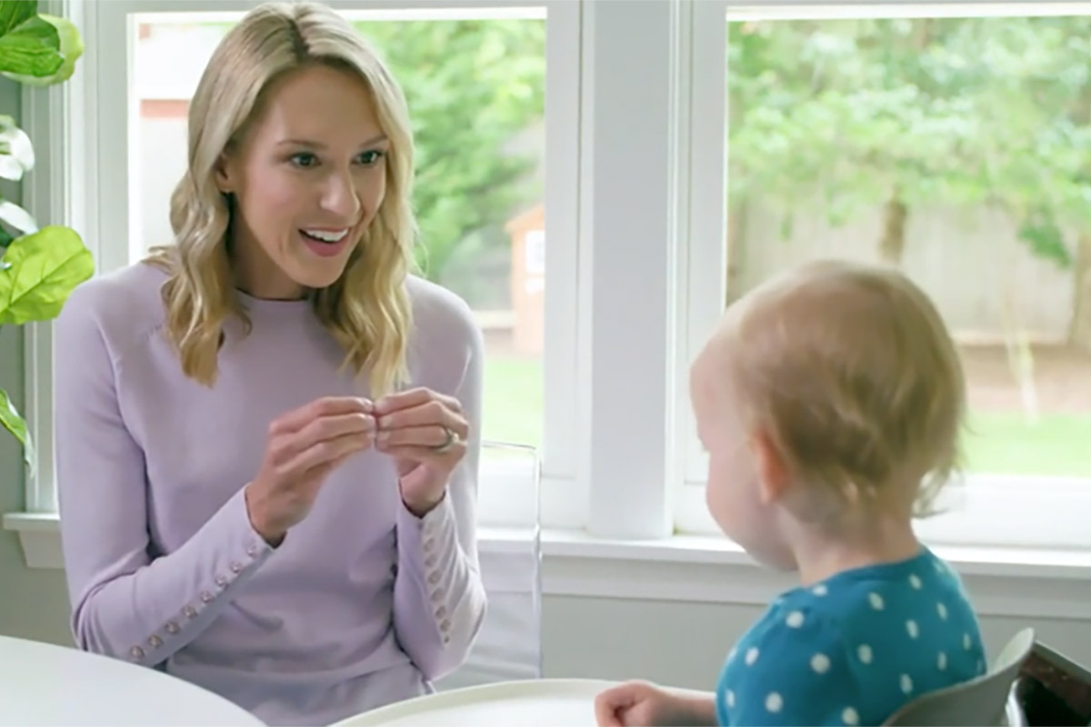 Woman sitting at a kitchen table doing the baby sign language sign for the word "more" while baby is watching