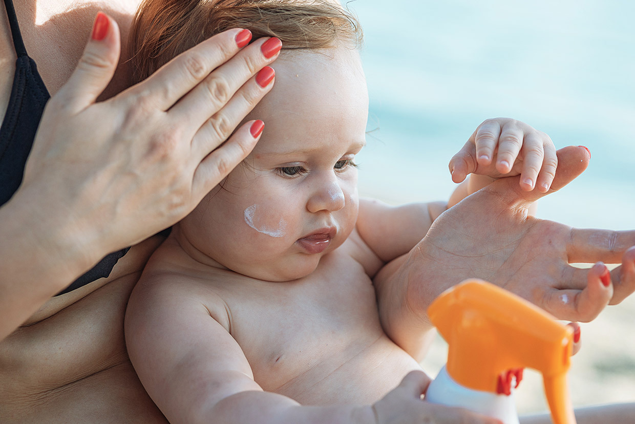 Baby sitting on parents lap at the beach, parent applies sunscreen to baby, baby holding sunscreen lotion
