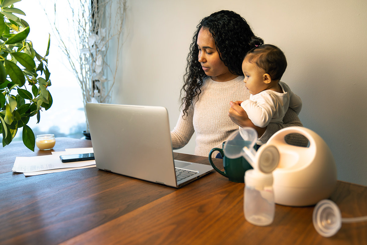 Parent holding their child with breast pump and bottles in the foreground, next to a laptop computer.