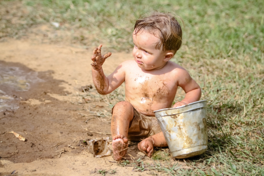 young toddler barefoot out on a lawn and covered in mud, holding a bucket