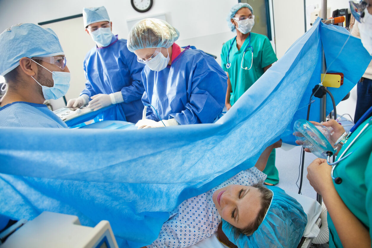 Woman during C-section surgery delivery wearing a surgical gown and hair net with blue drape in front of her, doctors are operating. 