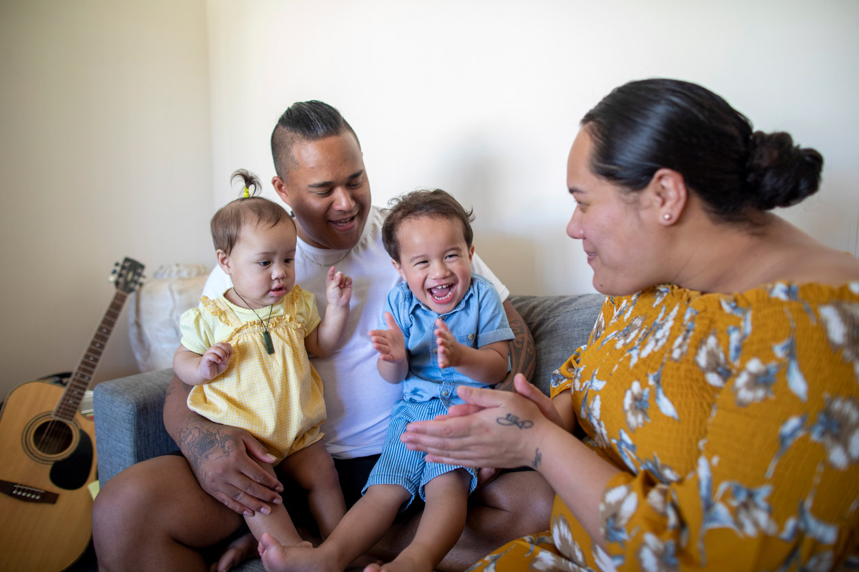 Parents sitting on couch with baby and toddler in their laps gesturing and signing together.