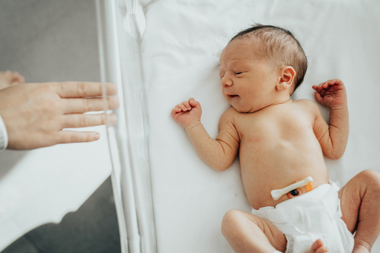 Newborn baby lying in a hospital bassinet, looking to the left, wearing only a diaper with umbilical cord clip visible. A parent's hand is reaching towards the bassinet from the left side.