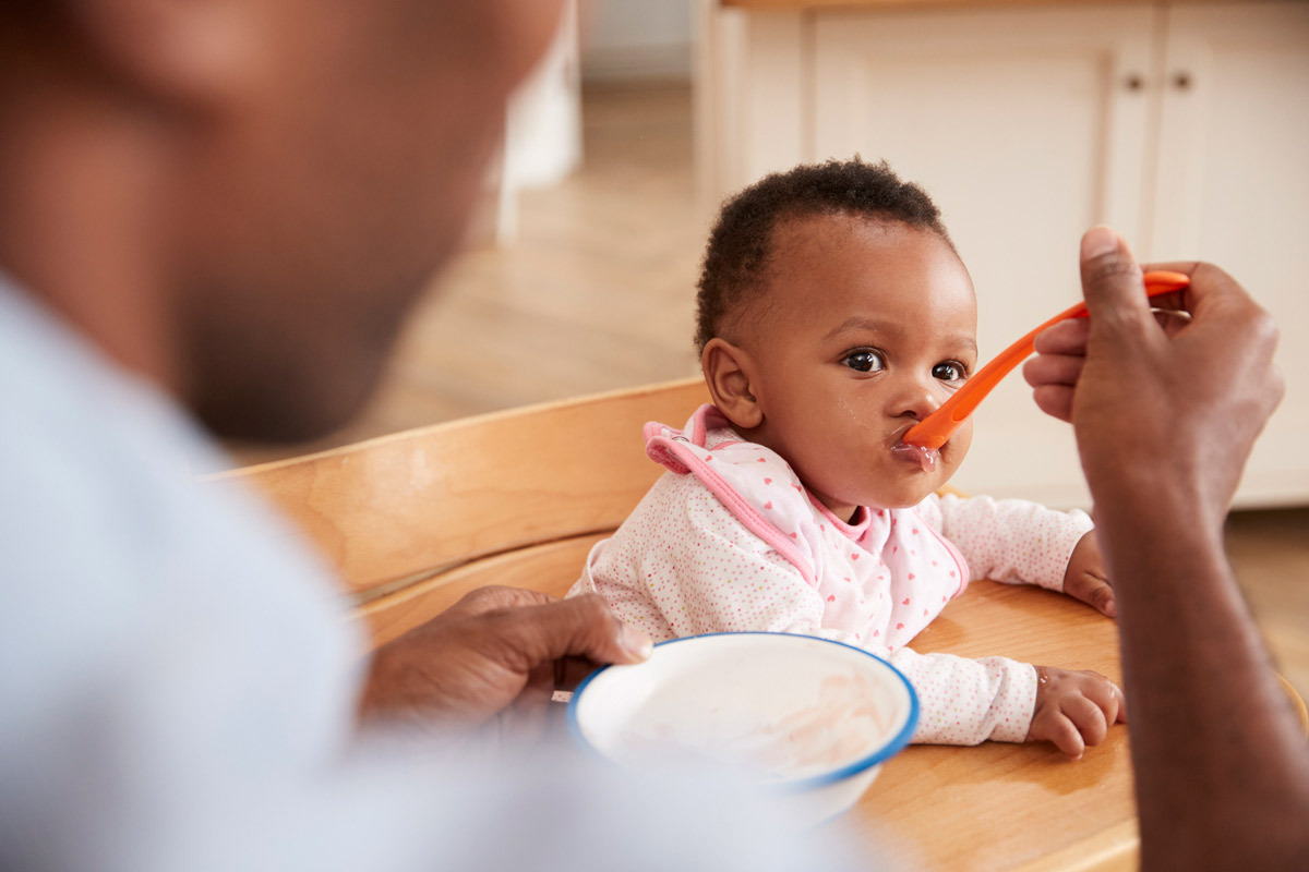 Person in the foreground spoon feeding pureed food to their infant baby who is sitting in a high chair. 