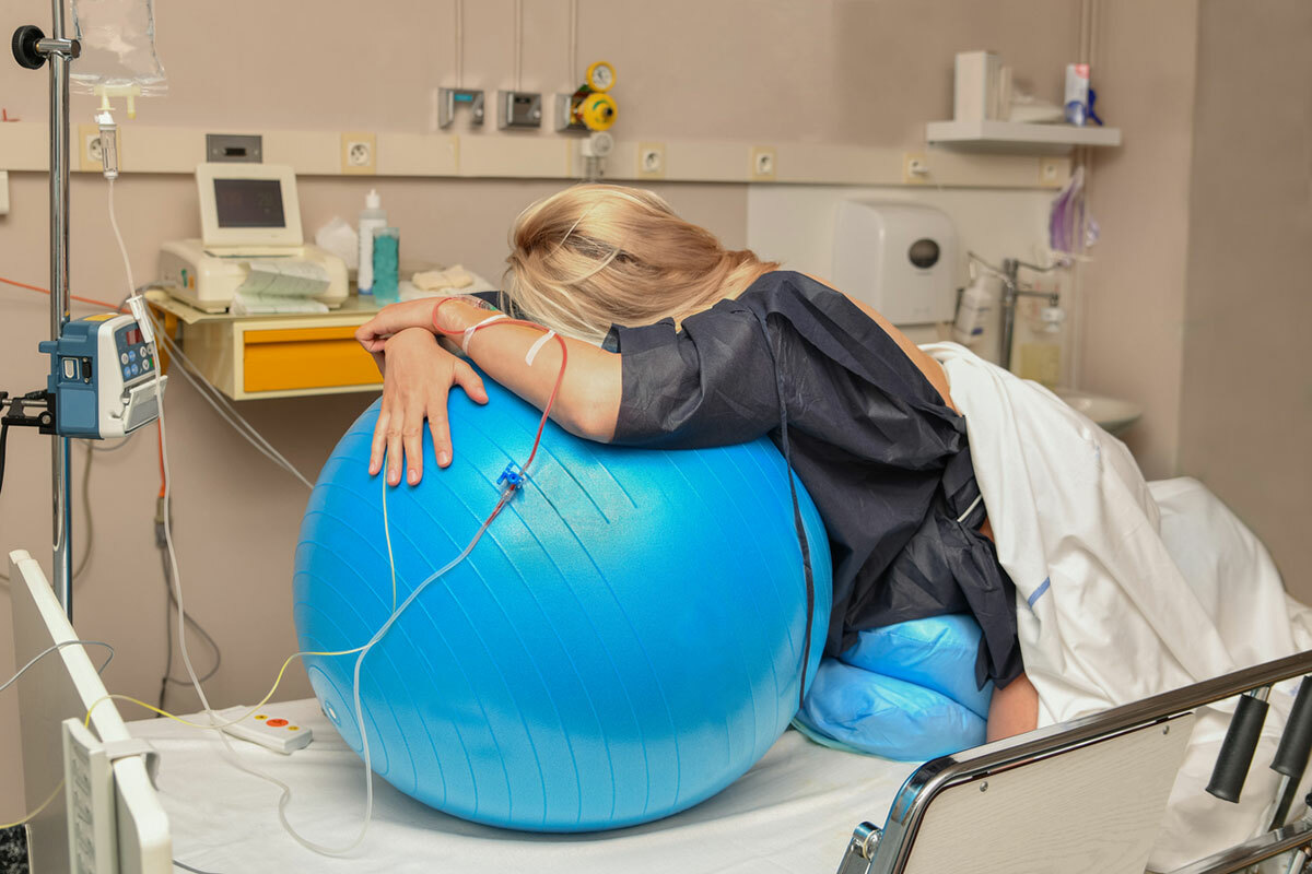 Blonde pregnant person during labor, kneeling on a hospital bed with their body draped over a blue physio birthing ball.