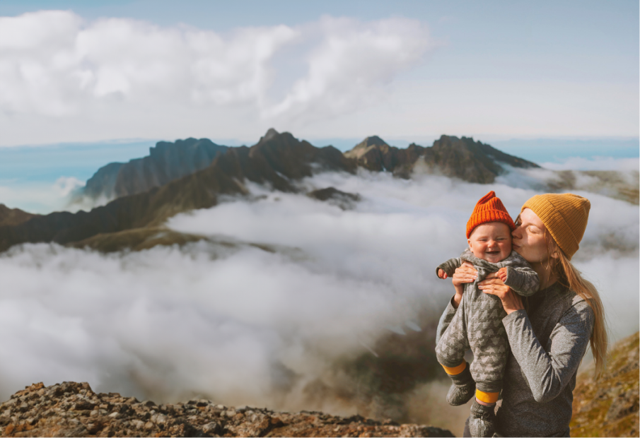 mother holding and kissing smiling baby on mountain range ridgeline with breathtaking foggy clouds in the background