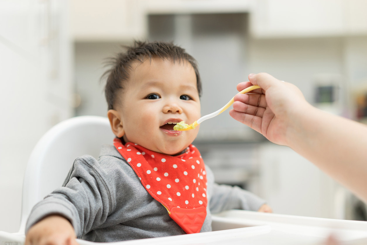Baby wearing a bib and sitting in a high chair, being spoon fed pureed food by an adult who is sitting out of the frame.