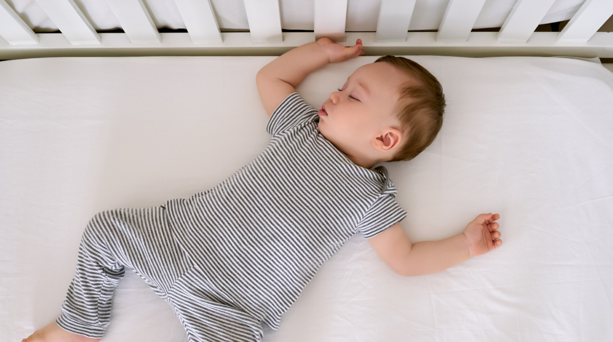 Overhead shot of an older baby wearing a striped romper sleeping soundly on their back in a white crib with a white sheet.