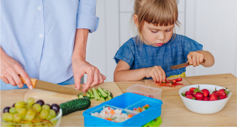 Girl chopping fruit on a cutting board, working with mother to pack her own lunch