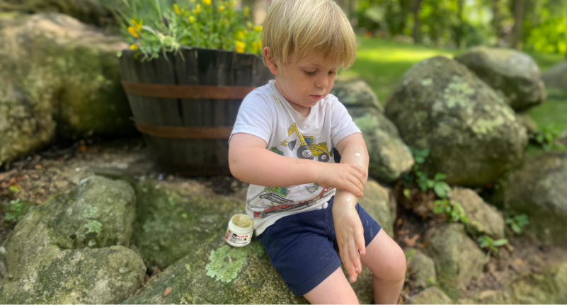 Toddler applying Sallyeander No-Bite-Me Cream to his arm, while sitting on a large rock outdoors